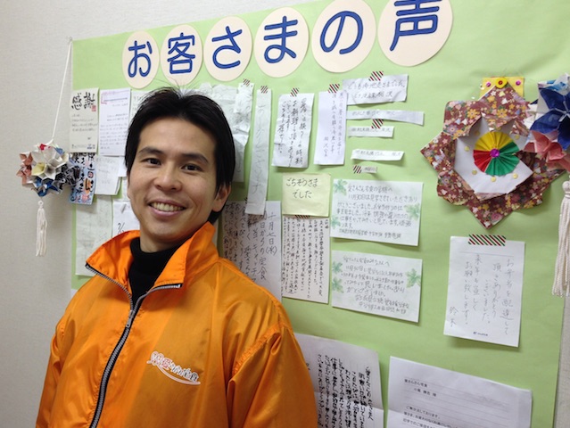 愛さんさん宅食株式会社代表の小尾勝吉さん。お客様の声が壁一面に掲示されている。感謝と喜びのメッセージだ。